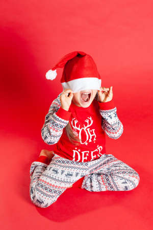 a happy girl in red Christmas pajamas with reindeer hides under a Santa Claus hat on a red backgroundの写真素材