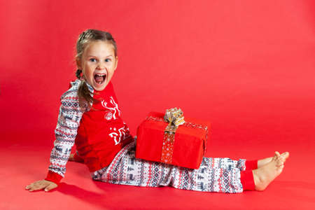 laughing girl with two pigtails and in Christmas pajamas sits on the floor and holds a gift box on her lapの写真素材