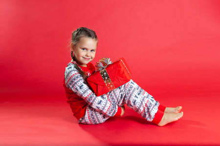 cute girl with two pigtails and in Christmas pajamas sitting on the floor, hugging her knees, holding a gift boxの写真素材