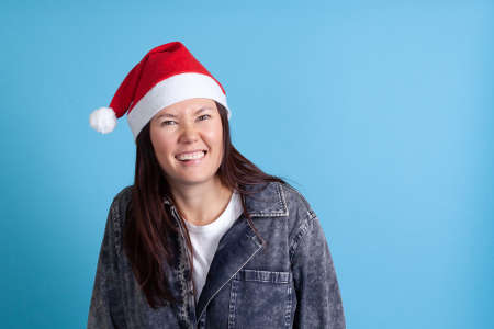 close up portrait of mischievous Asian young woman in Santa hat smiling and biting lip with her teeth on blue backgroundの写真素材