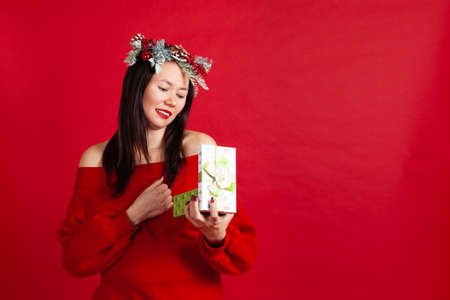 mock up of beautiful Asian young woman in Christmas wreath with red lipstick opens a new year gift, on a red backgroundの写真素材