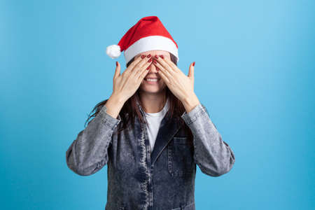 laughing Asian young woman in Santa Claus hat covers her eyes with hands waiting for gift or miracle, on blue backgroundの写真素材