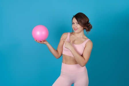 a slender woman in a pink Pilates suit holds a ball and points at it with her index finger, isolated on a blue backgroundの写真素材