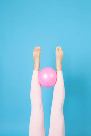 womens legs in pink sports leggings hold a rubber Pilates ball between them, isolated on a blue backgroundの写真素材