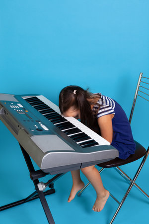 a crying girl sits at the piano and bows her head to the keys, hating the music school, isolated on a blue backgroundの写真素材