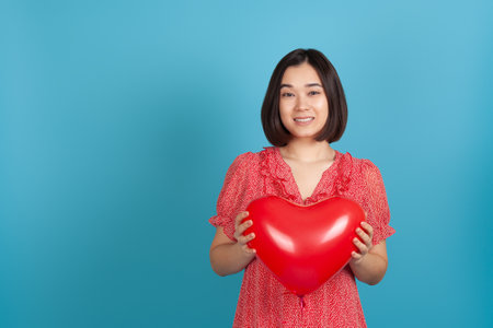 close up smiling happy Asian woman holding red heart shaped balloon on Valentines day isolated on blue backgroundの写真素材