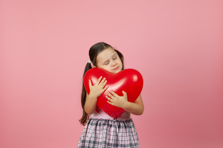 close-up gentle girl in pink dress with eyes closed hugs a red balloon in the shape of a heart to her head, isolated on a pink backgroundの写真素材