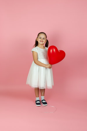 full-length careless, unconcerned, girl in white dress holding red heart shaped balloon isolated on pink backgroundの写真素材