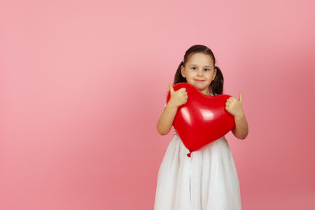 mock up kindly, hearted girl in white dress holding red balloon in the shape of heart, giving a thumbs up on two hands, isolated on a pink backgroundの写真素材