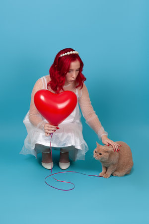 a young woman in a white dress holds a flying balloon in the shape of a heart, squats and strokes a red British cat, isolated on a blue backgroundの写真素材