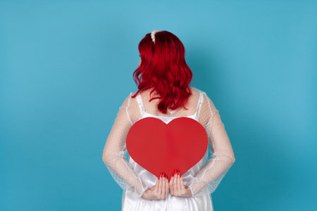 a young woman in a white dress and with red hair holds a large red paper heart behind her back hiding her face, isolated on a blue backgroundの写真素材