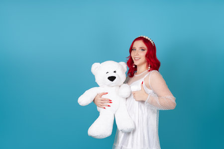 smiling happy young woman in white dress with red hair holds white teddy bear and gives thumbs up isolated on blue backgroundの写真素材