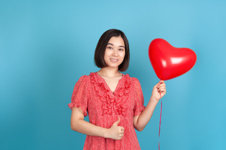 A smiling, joyful young Asian woman in a red dress holds a flying red heart-shaped balloon and gives a thumbs up isolated on a blue backgroundの写真素材