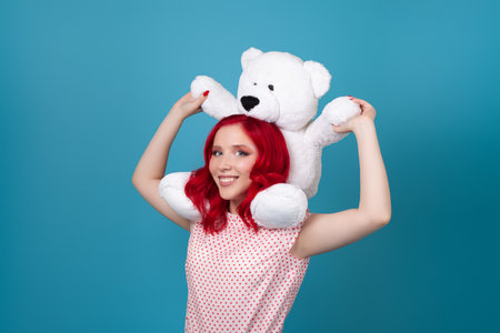 close up portrait of a laughing happy young woman with red hair and a huge teddy bear on her shoulders, isolated on a blue backgroundの写真素材