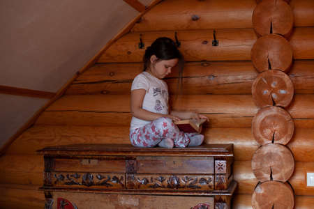 a thoughtful, serious girl sits cross-legged on an old wooden chest of drawers and reads a book in the bedroom of a rustic log houseの写真素材