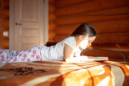 close-up a girl reading a book lies on her stomach in her pajamas on the bed in the bedroom of a log cabin in the villageの写真素材