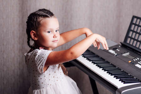 portrait of a mischievous, artistic girl in a white summer dress and two pigtails raising her hands to play the keys of an electronic synthesizer in a home interiorの写真素材