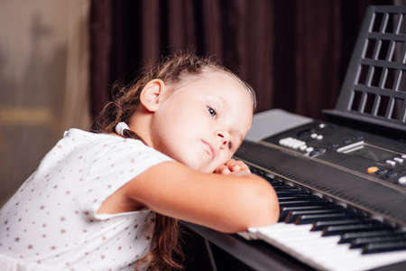 close-up portrait of a charming girl in a white dress and two pigtails lying with her head on the keys of an electronic synthesizer in a home interiorの写真素材
