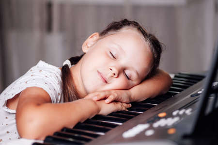close-up portrait of a sleeping girl with her eyes closed in a white dress and two pigtails lying with her head on the keys of an electronic synthesizer in a home interiorの写真素材