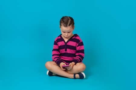 portrait of an enthusiastic girl in striped hoodie sitting cross-legged on the floor and playing games on a smartphone, isolated on a blue backgroundの写真素材