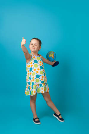 full-length portrait of a six-year-old girl in a white summer dress with colorful circles holding a globe in her hand and giving a thumbs upの写真素材