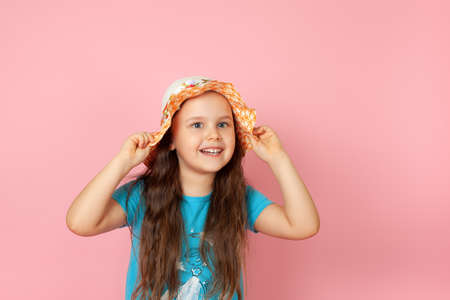 positive girl with long wavy hair in straw hat and blue dress looking sideways with her hands holding brim of her hat, isolated on a pink backgroundの写真素材