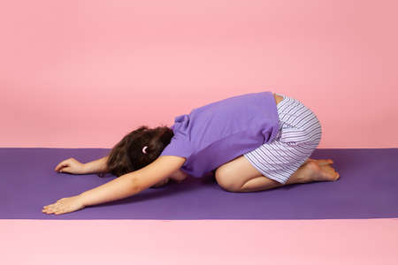 a girl does yoga on a purple mat in the pose of a child or Balasana, isolated on a pink backgroundの写真素材