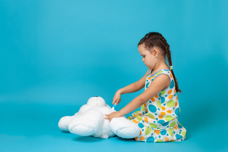 girl with pigtails sitting on the floor and playing surgeon performing scalpel surgery on a white teddy bear, isolated on a blue backgroundの写真素材
