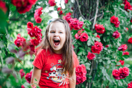 close-up portrait of a screaming, overjoyed girl with her mouth wide open in a red T-shirt near a rose bush in a park or gardenの写真素材