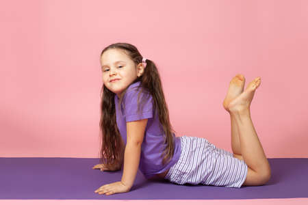 girl in profile doing morning exercises and stretching for the back muscles to improve flexibility on a purple mat, isolated on a pink backgroundの写真素材