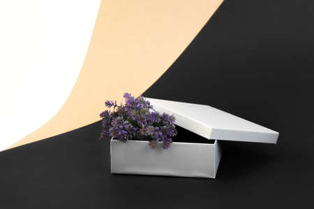open silver gift box with purple artificial flowers on a background of beige, black, white paper in...