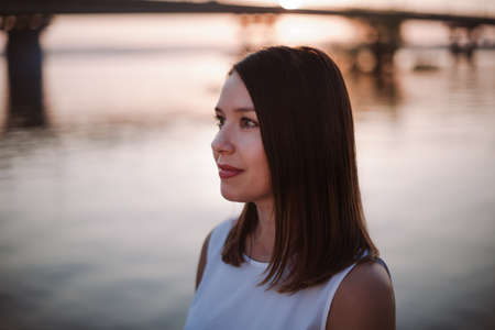 close-up portrait in profile of a young beautiful woman against the background of a river with a bridge on a summer evening at sunsetの写真素材
