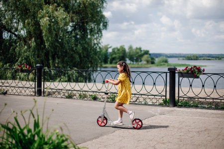lifestyle portrait a six-year-old girl in a yellow dress rides a scooter along the river on a sunny...