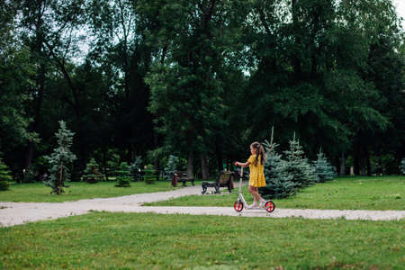summer postcard, a girl rides a scooter in a green summer park with trees and a green lawn on the backgroundの写真素材