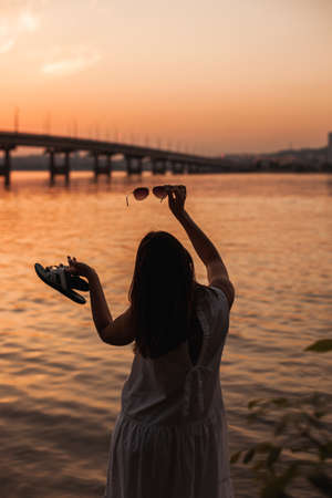 rear view of the silhouette of the back of a young woman holding sunglasses, slates in her outstretched hands and looking at the sunset and the bridge over the riverの写真素材
