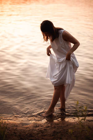 the silhouette of a young woman in a summer white dress walking along the edge of cool water on the beach by the river on a warm summer evening, an orange sunset and the orange surface of the riverの写真素材