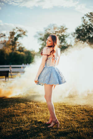 full-length portrait of a young woman in a short blue embroidered dress at a party in the park at sunset in summer with yellow smoke clouds on the backgroundの写真素材