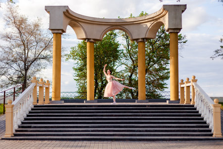 a ballerina dances with her leg raised in an arch of columns in urban architecture on the embankment of a river or sea on a sunny dayの写真素材