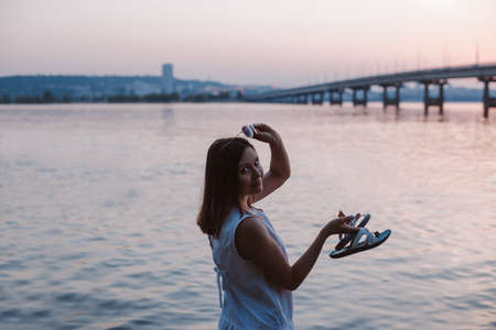 a slender brunette woman stands half-turned, takes off her sunglasses at sunset with a view of the...