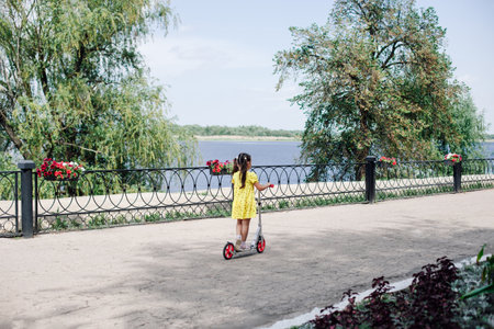 rear view of a girl in a yellow dress riding a scooter along the city embankment with flowers,...