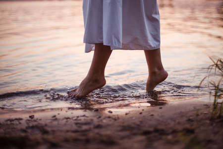 Womens feet in the river. Lifestyle close-up on the feet of a woman in a long white dress walking along the riverbank at sunsetの写真素材