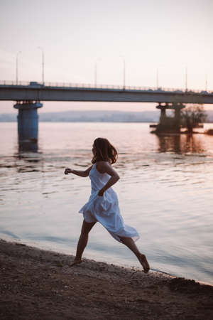 A woman runs along the river bank. Lifestyle portrait of a young woman in a white dress running fast barefoot on a sandy river bank with a bridge at sunset on a hot summer dayの写真素材