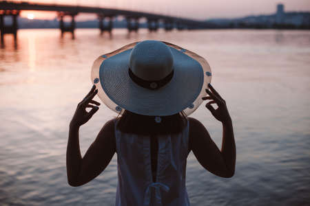 The silhouette of a woman by the river at sunset. Rear view of the silhouette of a woman in a straw...