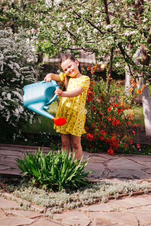 lifestyle full-length portrait of a smiling girl in a yellow dress watering flowers from a garden watering can, a girl with a watering can helps her parents in the gardenの写真素材