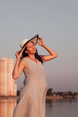 A pregnant woman at sunset. A peaceful, relaxed pregnant woman holds the brim of a straw hat with her hands and enjoys nature and inner harmonyの写真素材