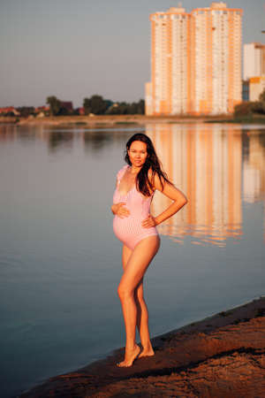 pregnant woman in a swimsuit, Lifestyle full-length portrait of a pregnant woman posing in a pink swimsuit holding her hands on a pregnant belly on a summer vacationの写真素材