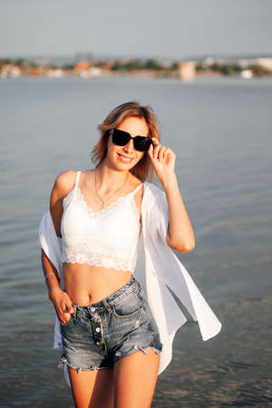 Photo of a woman in sunglasses. Portrait of a beautiful young woman in short denim shorts, a white top and a shirt with dark sunglasses on the background of water and landscape at dawnの写真素材