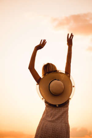 A young woman poses against the background of the sunset. Photo of a beautiful slender blonde with her hands raised up in a summer dress and a straw hat decorated with a ribbon and flowersの写真素材