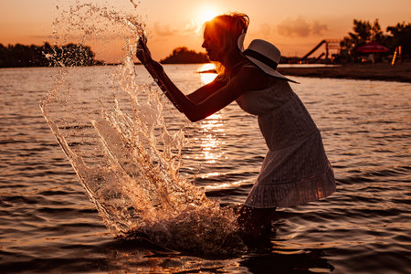 A young woman splashes water. Silhouette photo of a beautiful happy blonde in a summer dress and a straw hat with a ribbon on the background of the sunset and the beachの写真素材