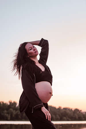Brunette poses against background of nature. Happy pregnant woman in dark silk unbuttoned pajamas and long brown hair fluttering in wind enjoys sunset against background of summer forestの写真素材